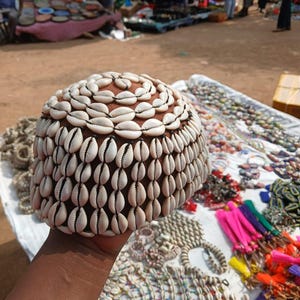 May include: A rounded cap covered in rows of white cowrie shells arranged in a circular pattern. The base material of the cap is a solid brown color, and the shells are attached with dark thread, creating a textured, layered appearance.
