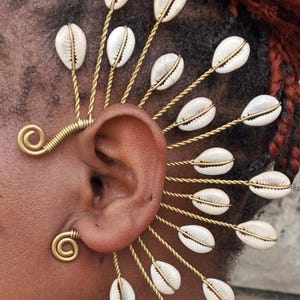 Pode incluir: Um ear cuff com conchas brancas e fio dourado. As conchas são dispostas em forma de leque, criando um acessório distinto. O ear cuff envolve a orelha, adicionando um toque boêmio. O design é intrincado.
