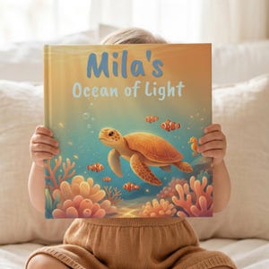 Puede incluir: Un libro infantil de tapa dura titulado Mila's Ocean of Light, con una ilustración de una tortuga marina nadando entre corales naranjas, pequeños peces payaso y un caballito de mar en una escena submarina azul y dorada.