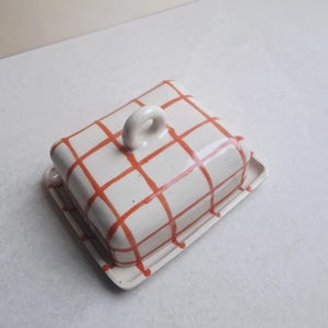Könnte beinhalten: Eine weiße Keramik-Butterdose mit einem rot karierten Muster auf dem Deckel. Der Deckel hat einen kleinen, abgerundeten Griff. Die Dose steht auf einer rechteckigen Basis.