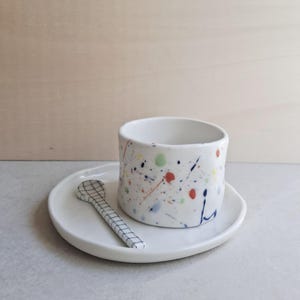 Könnte beinhalten: Ein Set aus einer weißen Keramiktasse und einer passenden Untertasse mit einem bunten Farbspritzermuster in Rot, Blau, Grün und Orange. Ein kleiner Keramiklöffel mit schwarzem Gittermuster liegt auf der Untertasse neben der Tasse.