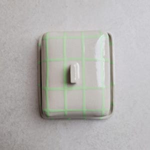 Könnte beinhalten: Eine quadratische Butterdose aus Keramik mit Deckel. Die Dose ist cremefarben mit einem hellgrünen Gittermuster. Ein kleiner, rechteckiger Griff befindet sich auf dem Deckel. Der Hintergrund ist hellgrau.