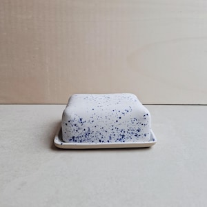 Può includere: Una burriera in ceramica bianca con coperchio, macchiata di vernice blu. Il piatto rettangolare ha angoli arrotondati e una base leggermente rialzata. Il coperchio presenta un motivo maculato abbinato.