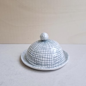 Puede incluir: Una mantequillera de cerámica blanca con una tapa en forma de cúpula que presenta un patrón de cuadrícula negra dibujado a mano. La tapa tiene un pequeño pomo esférico y la base es un plato circular a juego con un borde ligeramente elevado.