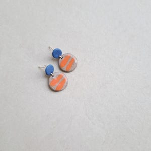 May include: A pair of ceramic earrings with a light gray base, orange stripes, and a small blue circle at the top.
