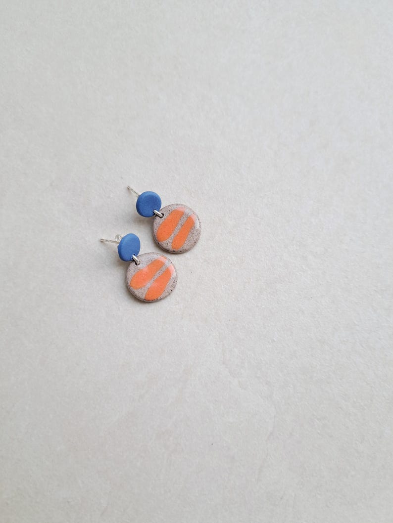 May include: A pair of ceramic earrings with a light gray base, orange stripes, and a small blue circle at the top.