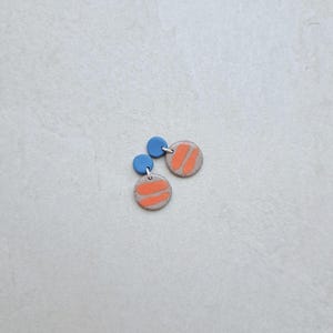 May include: A pair of small, round earrings with a light gray base and a bright orange stripe design. The earrings have a small blue circle at the top.