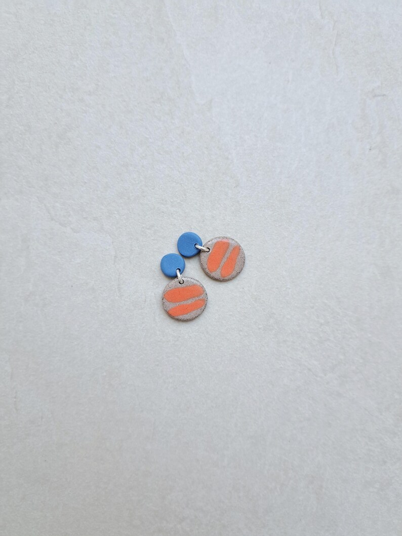 May include: A pair of small, round earrings with a light gray base and a bright orange stripe design. The earrings have a small blue circle at the top.