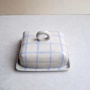 Op de afbeelding: Een keramische botervloot met een deksel en een dienblad. De schaal is wit met een blauw ruitpatroon. Het deksel heeft een lusvormige handgreep. De botervloot is rechthoekig en staat op een wit oppervlak.