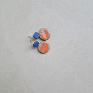May include: A pair of ceramic earrings with orange and white geometric designs. The earrings have blue accents.