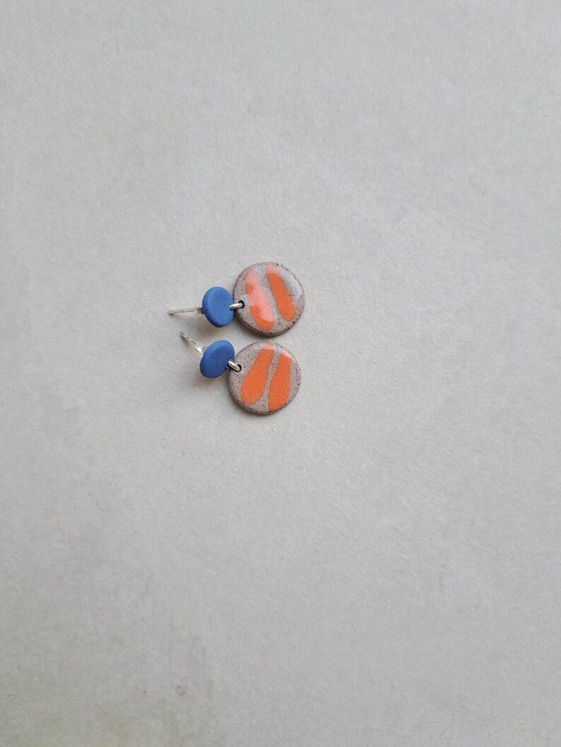 May include: A pair of ceramic earrings with orange and white geometric designs. The earrings have blue accents.