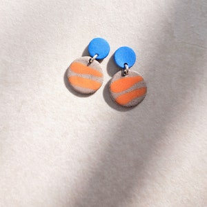 May include: A pair of dangle earrings with a round, orange and gray patterned design and a small, blue circle at the top.