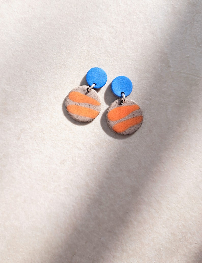May include: A pair of dangle earrings with a round, orange and gray patterned design and a small, blue circle at the top.