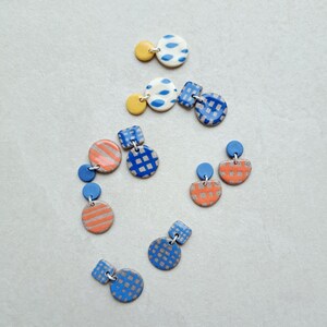 May include: A set of six pairs of handmade ceramic earrings. The earrings are in various geometric shapes and feature different patterns in blue, yellow, and orange.