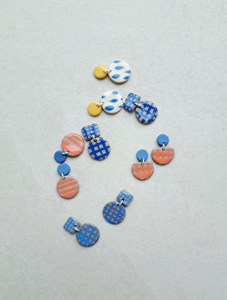 May include: A set of six pairs of handmade ceramic earrings. The earrings are in various geometric shapes and feature different patterns in blue, yellow, and orange.