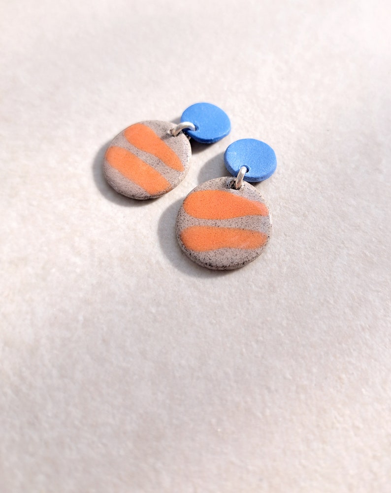 May include: A pair of handmade ceramic earrings with orange and gray striped circles and blue round accents.
