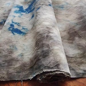May include: A rolled-up piece of fabric with a textured, abstract design. The fabric features a blend of gray, blue, and white hues, creating a watercolor effect. The material appears to be a heavy weave, suitable for upholstery or home decor.