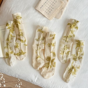 May include: Three pairs of sheer, off-white crew socks featuring a botanical print of green leaves and stems. The socks are arranged on a white fabric surface alongside a small paper calendar and a decorative branch with white berries.