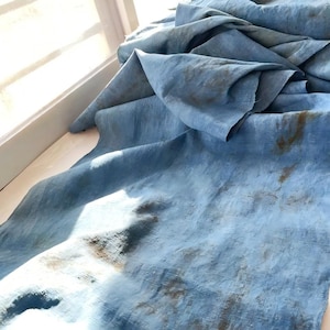Può includere: Un grande pezzo di tessuto blu, forse lino o cotone, con macchie marroni, è drappeggiato vicino a una finestra. Il tessuto ha un aspetto strutturato ed è piegato in più strati, creando ombre e luci.