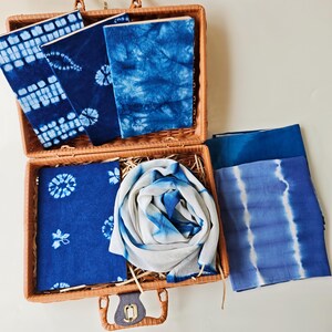Peut inclure: Une valise en osier tressé contenant plusieurs articles en tissu arborant des motifs tie-dye bleu et blanc de style shibori. La collection comprend des pièces de tissu rectangulaires, une écharpe pliée et des carnets recouverts de tissu indigo.