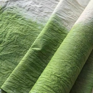 May include: Close-up of a textured fabric with a gradient of white to green. The fabric is folded and rolled, showcasing the color transition. The material appears to be lightweight and slightly crinkled, suggesting a soft texture. Ideal for textile projects.