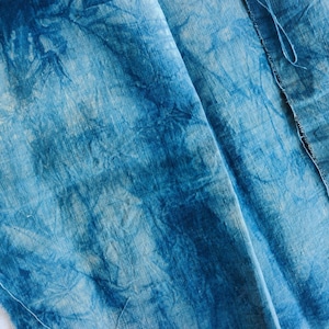 May include: Close-up of a piece of blue fabric with a tie-dye pattern. The fabric has a textured appearance with varying shades of blue, creating a marbled effect. The edges of the fabric are visible, showing the raw, unfinished edges.