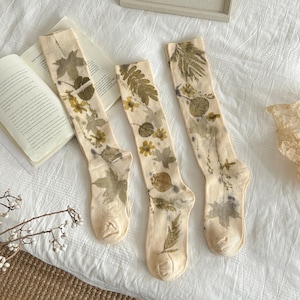 May include: Three off-white crew socks featuring a botanical print of various pressed green and yellow leaves and small flowers. The socks are arranged vertically, displaying a detailed pattern of foliage that covers the length of the fabric.