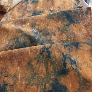 May include: Close-up of a folded textile with a mottled pattern. The fabric features a blend of brown and orange hues, with darker blue-green accents creating a marbled effect. The texture appears slightly wrinkled, suggesting a natural fiber.
