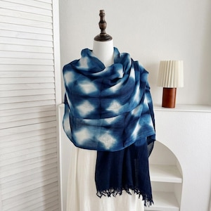 Peut inclure: Écharpe tie-dye bleue et blanche drapée sur un mannequin. L'écharpe a une bordure bleu foncé et un bord effiloché. Le motif est une série de formes de losanges blancs sur fond bleu. L'écharpe est présentée sur un mannequin blanc.