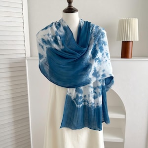 Könnte beinhalten: Ein leichter Schal mit einem Batikmuster in tiefen Blau- und Weißtönen. Der Stoff hat eine geknitterte, strukturierte Optik mit weichen, ausgefransten Kanten und ist elegant über eine Schaufensterpuppe mit schlichtem, weißem Kleidungsstück drapiert.