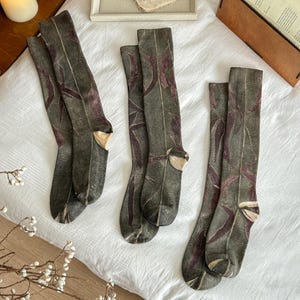 May include: Three pairs of knee-high socks in a muted olive green color. Each sock features a repeating pattern of dark purple and maroon leaf silhouettes. The socks have a visible seam running down the center of the foot and a light beige heel.