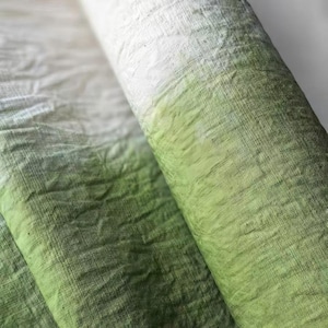 Puede incluir: Un rollo de tela con un degradado de colores, que va del blanco a tonos verdes. La tela tiene un aspecto texturizado, con hilos tejidos visibles y ligeras arrugas. El rollo está parcialmente desenrollado, revelando más tela verde.