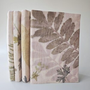 May include: A set of four rectangular books with fabric covers in a light pink hue. Each cover features botanical imprints of various leaves and small plants in shades of brown, olive green, and yellow, creating a natural, organic pattern.