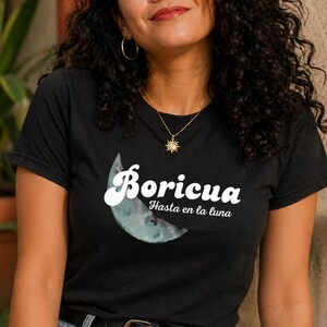May include: Black t-shirt with the word "Boricua" in white script above a watercolor-style crescent moon. Below the moon, the text "Hasta en la luna" is printed in white. The model is wearing blue jeans and a black belt.