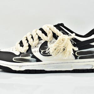 Peut inclure: Sneaker personnalisée noire et blanche avec des lacets en corde et une pampille. La chaussure présente une semelle noire, une semelle intermédiaire blanche et des détails peints en noir et blanc. Le texte "WAVE" et "CUSTOM" est visible.