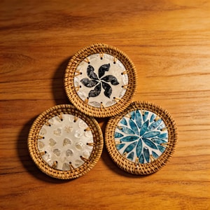 May include: Three round coasters with woven rattan edges. Each coaster features a different design: a black floral pattern, a white heart pattern, and a blue floral pattern. The coasters are set on a wooden surface.