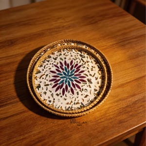 May include: Two round woven trays nested together. The trays have a natural brown color with a decorative floral design in the center. The floral design is composed of burgundy and teal petals on a white background.