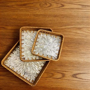 Puede incluir: Tres bandejas cuadradas de ratán tejido con incrustaciones de nácar. Las bandejas están dispuestas sobre una superficie de madera. La incrustación de nácar tiene un diseño floral. Las bandejas tienen un borde cosido y el texto "MOC AN Home".