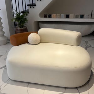 May include: A modern, off-white, oval-shaped ottoman with a matching bolster pillow and a small, textured, spherical pillow. A brown bolster pillow is also present. The furniture is set on a white marble-patterned floor.