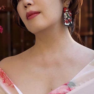 May include: A rectangular drop earring featuring a graphic illustration of a small green and yellow bird perched on a dark tree branch against a white background with red floral accents, suspended from a small gold-toned metal circular stud.