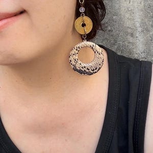以下が含まれることがあります： 小さな透明なビーズ、中央に穴の開いた円形のゴールドトーンのコイン、そしてベージュと黒のヘビ革模様のテクスチャー仕上げが施された大きな円形のペンダントが、金属製のリンクチェーンからぶら下がっているドロップイヤリング。