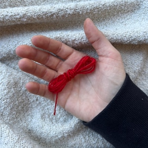 May include: A small bundle of bright red yarn rests in an open hand. The yarn is tightly wound, with a single strand extending downwards. The background is a textured, light-colored fabric, and the person is wearing a black long-sleeved shirt.