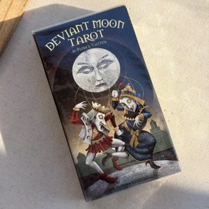 Può includere: Un mazzo di tarocchi intitolato "Deviant Moon Tarot" di Patrick Valenza. La scatola presenta uno sfondo blu scuro con un grande volto bianco a forma di luna. Due figure in costume sono raffigurate sul davanti, con le parole "U.S. GAMES SYSTEMS, INC." in basso.