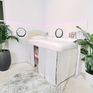 Könnte beinhalten: Ein weißer Massagetisch mit cremefarbenem Kopfteil und weißem Bezug. Der Tisch steht in einem weißen Raum mit zwei schwarzen kreisförmigen Spiegeln und Topfpflanzen. Auf dem Boden liegt ein Teppich.