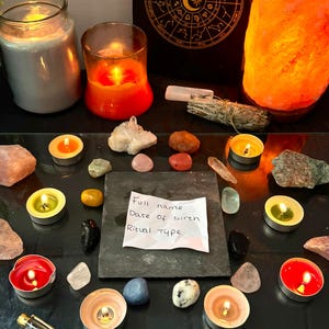 May include: A square slate tile with a white paper note reading Full name, Date of birth, Ritual type, surrounded by various crystals, tea light candles in red, orange, and green, a sage bundle, a salt lamp, and a gold pen on a glass surface.