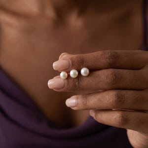 May include: Three white pearl stud earrings are held between fingers. The pearls are round and have a lustrous sheen. The earrings are set on gold posts. The background is a blurred image of a person wearing a purple top.