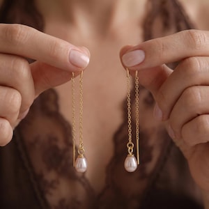 May include: A pair of gold threader earrings featuring delicate fine chains and a single teardrop-shaped pearl pendant suspended from each end. The earrings are constructed with a straight gold post that connects to the chain, allowing the jewellery to hang vertically with a polished finish.