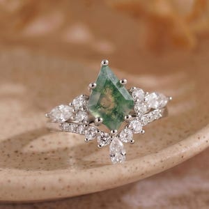 May include: A silver-toned ring featuring a central kite-shaped green moss agate stone. The band is adorned with a cluster of small, clear, round and marquise-cut stones arranged in a V-shape, extending along the sides of the central gemstone.