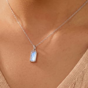 May include: A silver-toned necklace featuring a rectangular pendant with a translucent, iridescent moonstone-like gem. The pendant is suspended from a delicate, thin silver-toned chain that rests against the collarbone area of a beige ribbed top.
