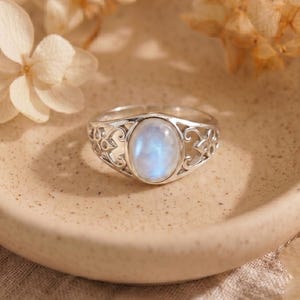 May include: A silver ring featuring an oval-shaped, iridescent moonstone cabochon set in a decorative metal band with intricate cut-out scrollwork patterns. The gemstone displays a soft blue glow against the polished silver metal setting.